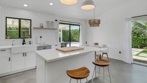 A modern kitchen with white cabinetry, a large island, pendant lighting, and a view of greenery outside. - Desert Oasis w/ Pool & Spa, Game Room + Theater (Indio)