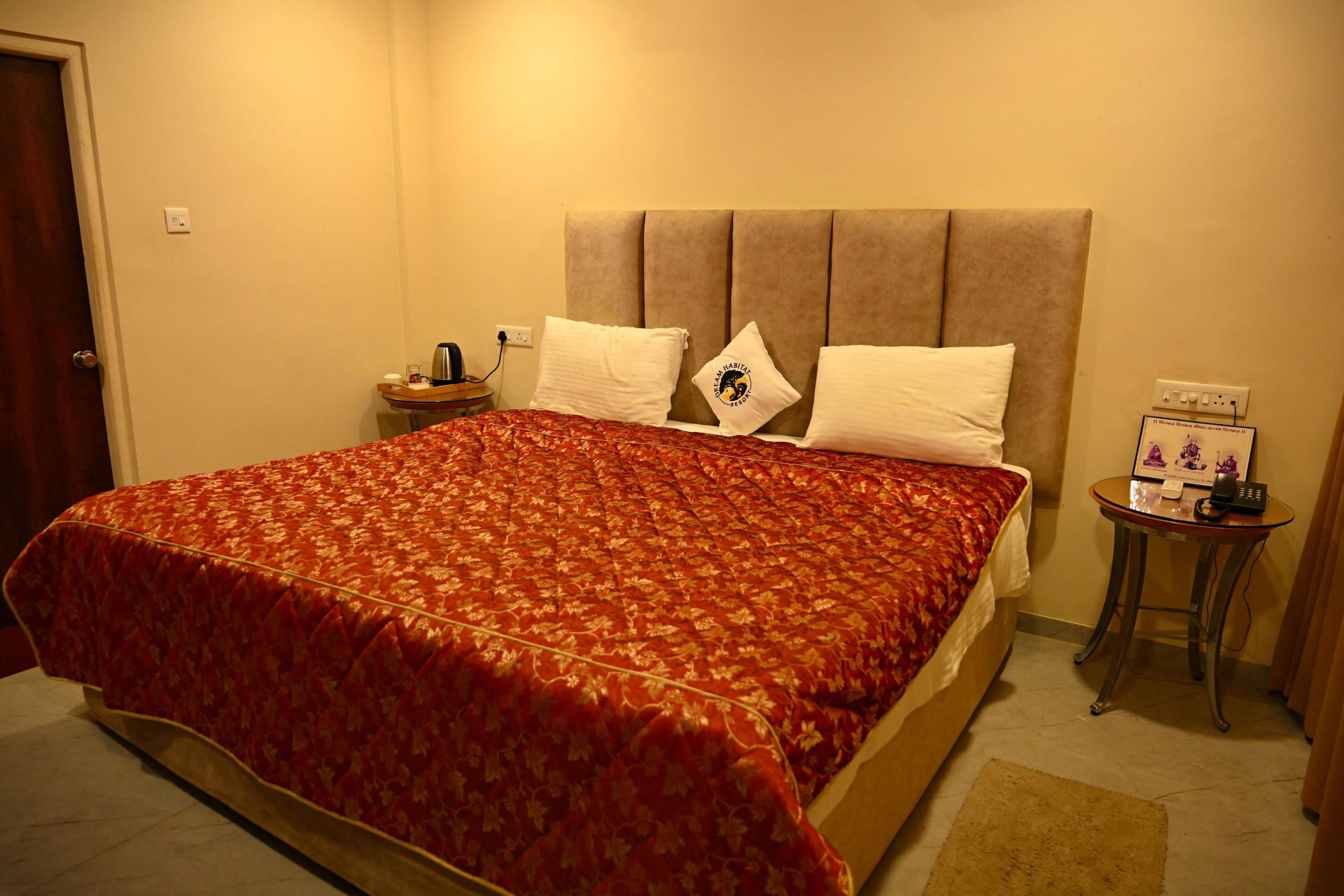 A hotel room with a bed, a bedside table with a lamp, a small table with a phone and a picture frame, and a door.