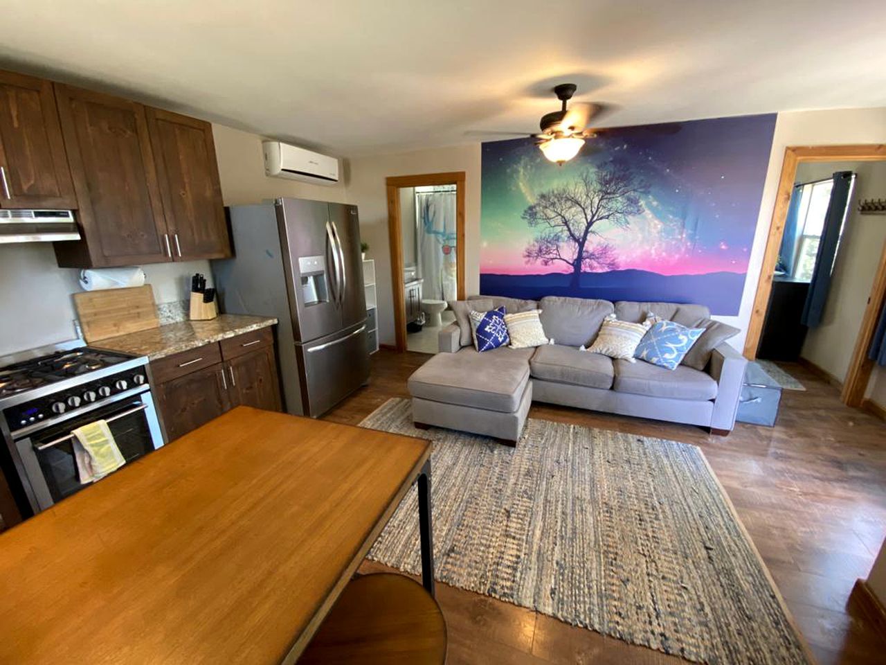 A modern kitchen with a dining area, a gray sofa, and a large painting of a tree.