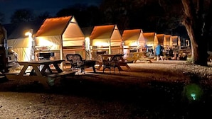 Varias tiendas de campaña con mesas y sillas, montadas en un camping durante la noche.