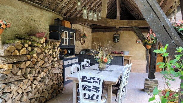 A rustic outdoor dining area with a stone wall, stacked firewood, and a table with chairs.