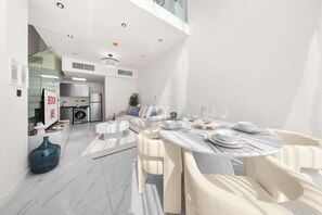 A modern kitchen with white cabinets, a washing machine, and a dining area with a round table set for four. - Free housekeeping visits during your stay - StayShort - Cozy 1BR Townhouse loft style located in Rukan Community (Dubai Land)