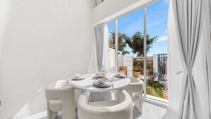 Uma área de refeições com uma mesa redonda e quatro cadeiras, janelas amplas com cortinas, e vista para um jardim.