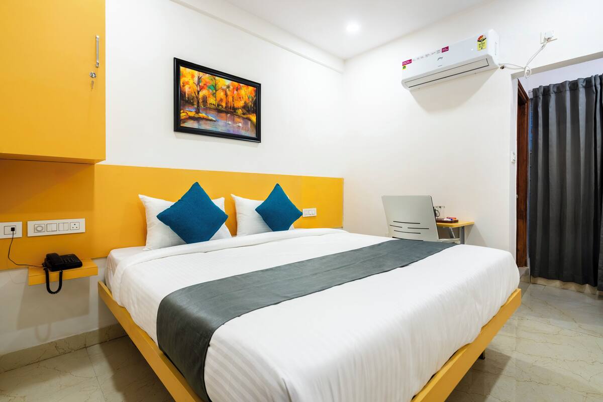 Una habitación de hotel con cama, mesita de noche, unidad de aire acondicionado, un cuadro en la pared y una cortina.
