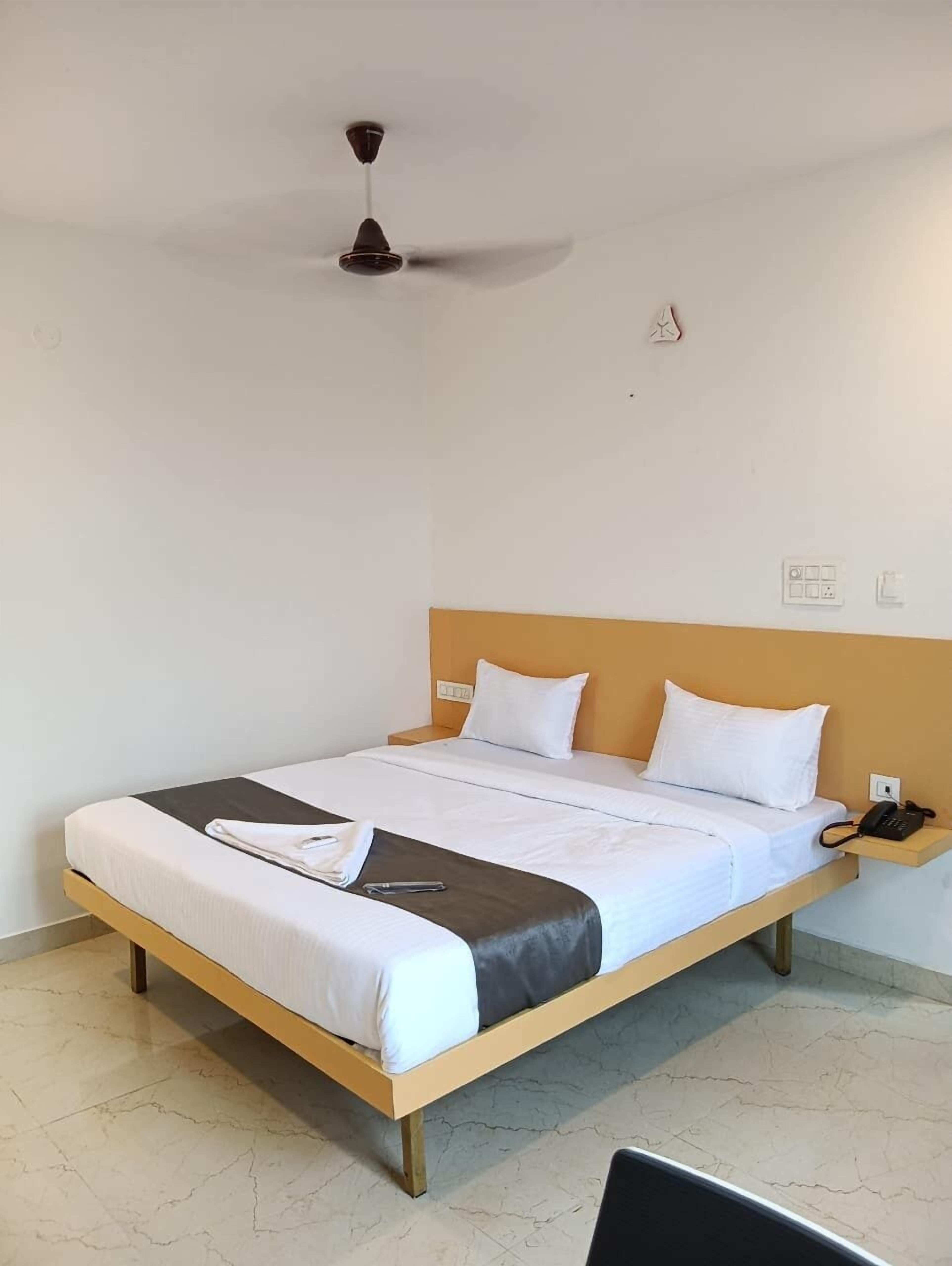A hotel room with a bed, a bedside table with a phone, a ceiling fan, and a wall with a power outlet.