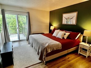 Un dormitorio con cama, mesita de noche, lámpara, un cuadro en la pared y vistas a través de un ventanal corredero.