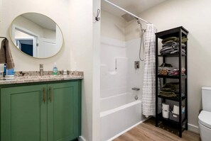 A bathroom with a green vanity, a round mirror, a shower with a white curtain, and a black storage unit with towels.