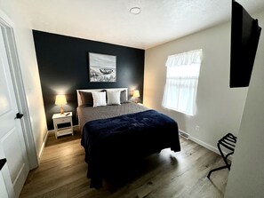 Un dormitorio con cama, mesitas de noche, lámpara, ventana y televisor montado en la pared.