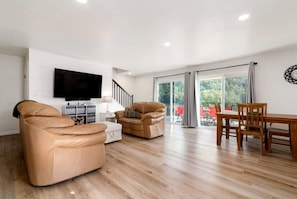 A spacious living area with a flat-screen TV, leather sofas, a wooden dining table, and a staircase leading to a balcony with outdoor seating.