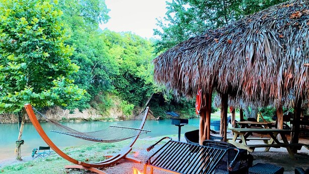 A hammock, a grill, and a thatched-roof structure by a river. - Incredible Glamping Retreat near Kingsbury, Texas: Luxury Safari Tent on the San Marcos River Perfect for a Family (Kingsbury)