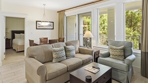 Ein Wohnzimmer mit einer Couch, einem Sessel und einem Couchtisch. Es gibt einen Deckenventilator, eine Lampe und ein Fenster mit Blick auf Grünflächen.