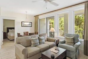 Ein Wohnzimmer mit einer Couch, einem Sessel und einem Couchtisch. Es gibt einen Deckenventilator, eine Lampe und ein Fenster mit Blick auf Grünflächen.