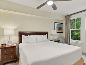 A hotel room with a large bed, a bedside table with a lamp, a nightstand with a clock, a ceiling fan, and a window with curtains. - One Bedroom Condo in Lasata (Miramar Beach)