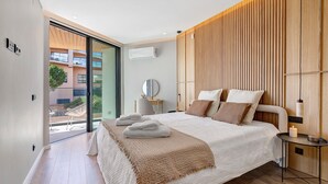 A modern bedroom with a large bed, a bedside table with a lamp, a view through a sliding door to an outdoor area, and a wooden panel wall. - Ria Front Paradise – A Gem Facing the Ria (Olhão)