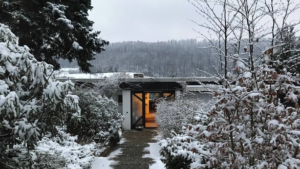 Un sendero cubierto de nieve que lleva a un edificio con puerta de vidrio, rodeado de árboles cargados de nieve y un fondo boscoso.