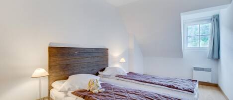 Una habitación con dos camas, una mesita de noche con lámpara, un cabecero de madera y una ventana con cortina.