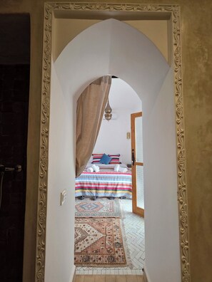 Ein rundbogenförmiger Durchgang führt in ein Schlafzimmer mit einer gestreiften Bettdecke, einem gemusterten Teppich und einer hängenden Leuchte.