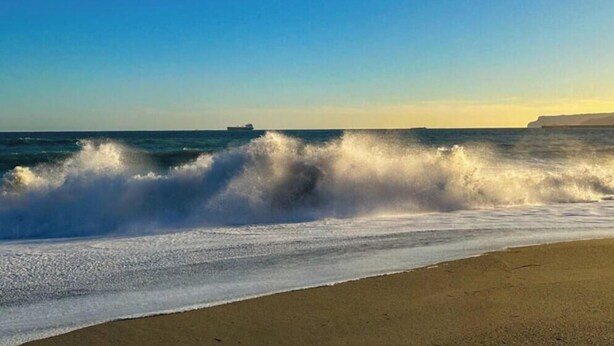 A beach with waves crashing onto the shore, a distant ship, and a cliff in the background.