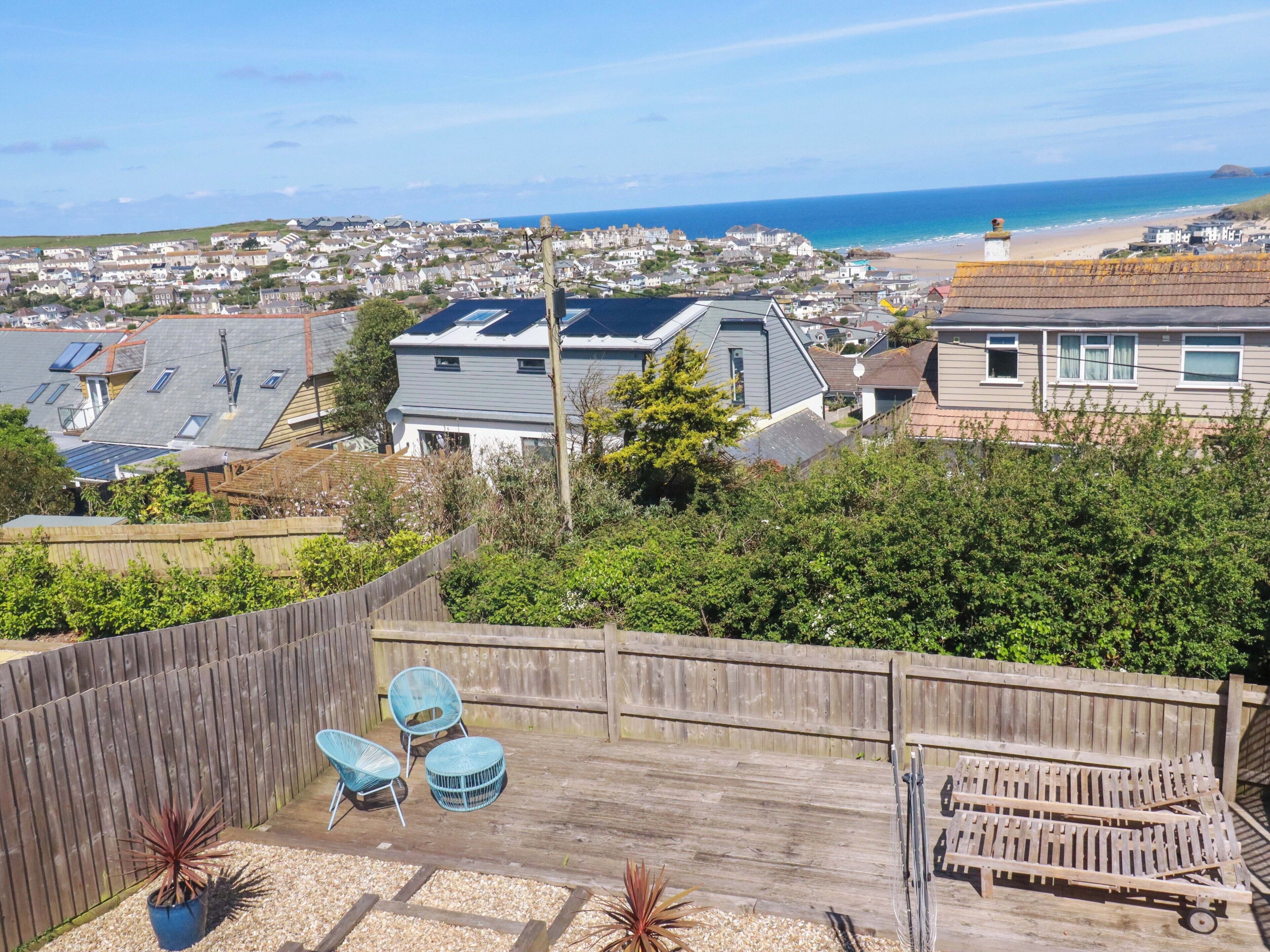 A backyard with a wooden deck, two blue chairs, a small table, and a sun lounger.