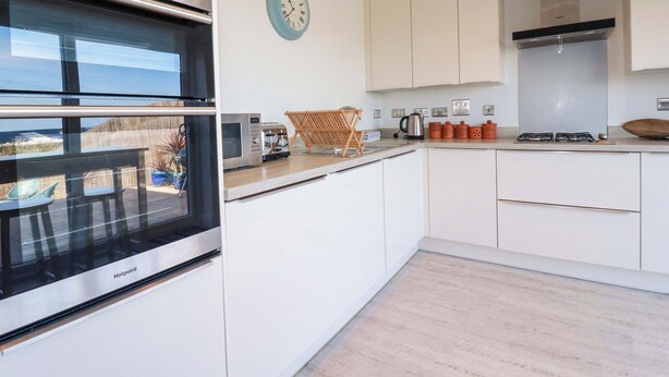 A modern kitchen with white cabinetry, a built-in oven, a coffee maker, and a clock on the wall. - Serfaus (Perranporth)