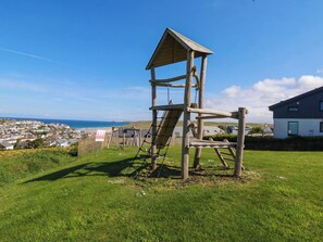 Eine hölzerne Spielkonstruktion mit Rutsche und Kletterbereich, eingebettet in eine grüne Wiese mit Blick auf eine Küstenstadt und Häuser im Hintergrund.