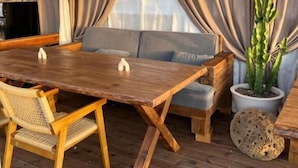 Una mesa de comedor de madera con sillas, un sofá, un televisor y una planta en maceta en una habitación con suelo de madera y cortinas.