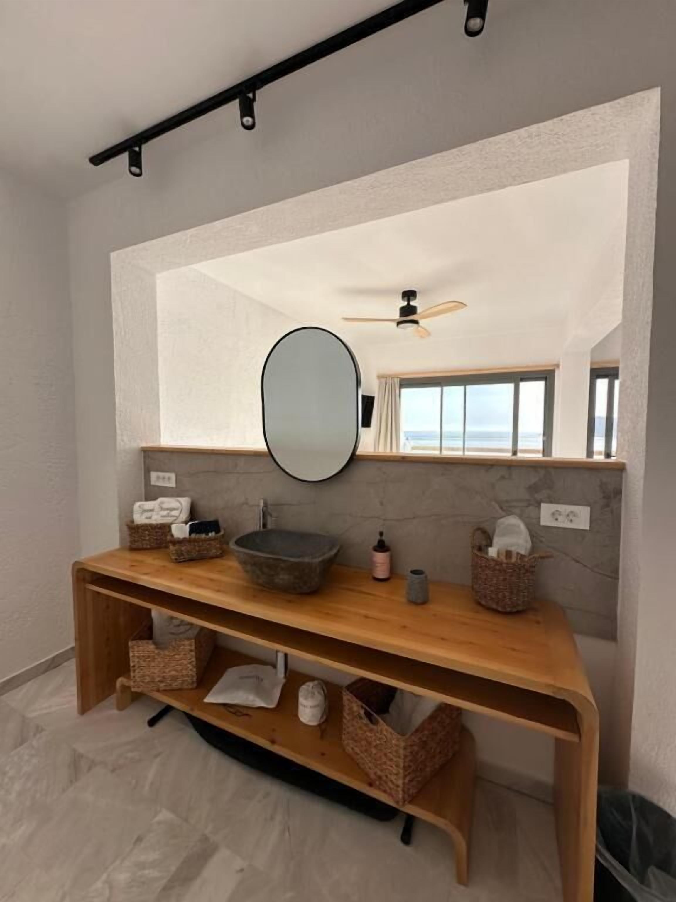 Un baño con tocador de madera, un espejo grande, un ventilador de techo y vistas al océano a través de amplios ventanales.