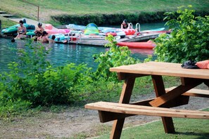 Une table de pique-nique en bois, près d’une étendue d’eau calme où l’on peut voir des bateaux et des bouées gonflables.