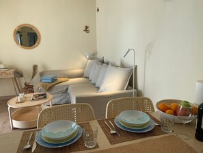 A living room with a sofa, a round mirror, a small table with a vase and candles, and a dining area with plates, cutlery, and a bowl of fruit. - Vacation Home 'Apartment Colores De Fuerteventura2' with Balcony and Wi-Fi (Costa Calma)