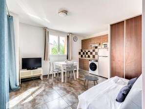 A compact hotel room with a bed, a TV, a dining area, a washing machine, and a kitchenette. - Tranquil Village Apartment for 6 (Saint-Cyprien)