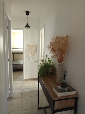 Ein Flur mit einem Bett im Hintergrund, einer Pendelleuchte, einem Holztisch mit einer Pflanze, einer Vase und einem Buch.