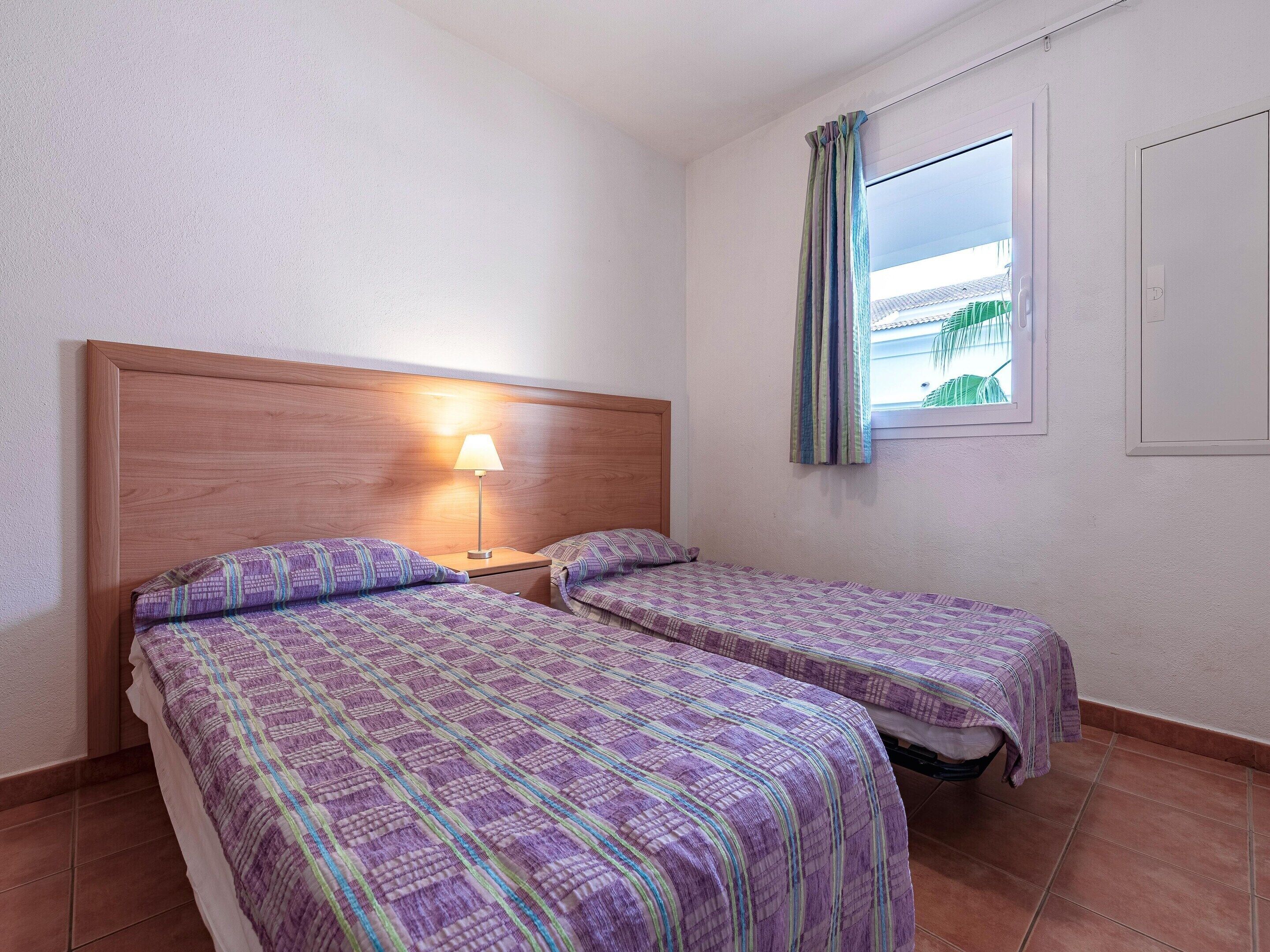 A room with two beds, a nightstand with a lamp, a window with curtains, and a view of greenery outside.