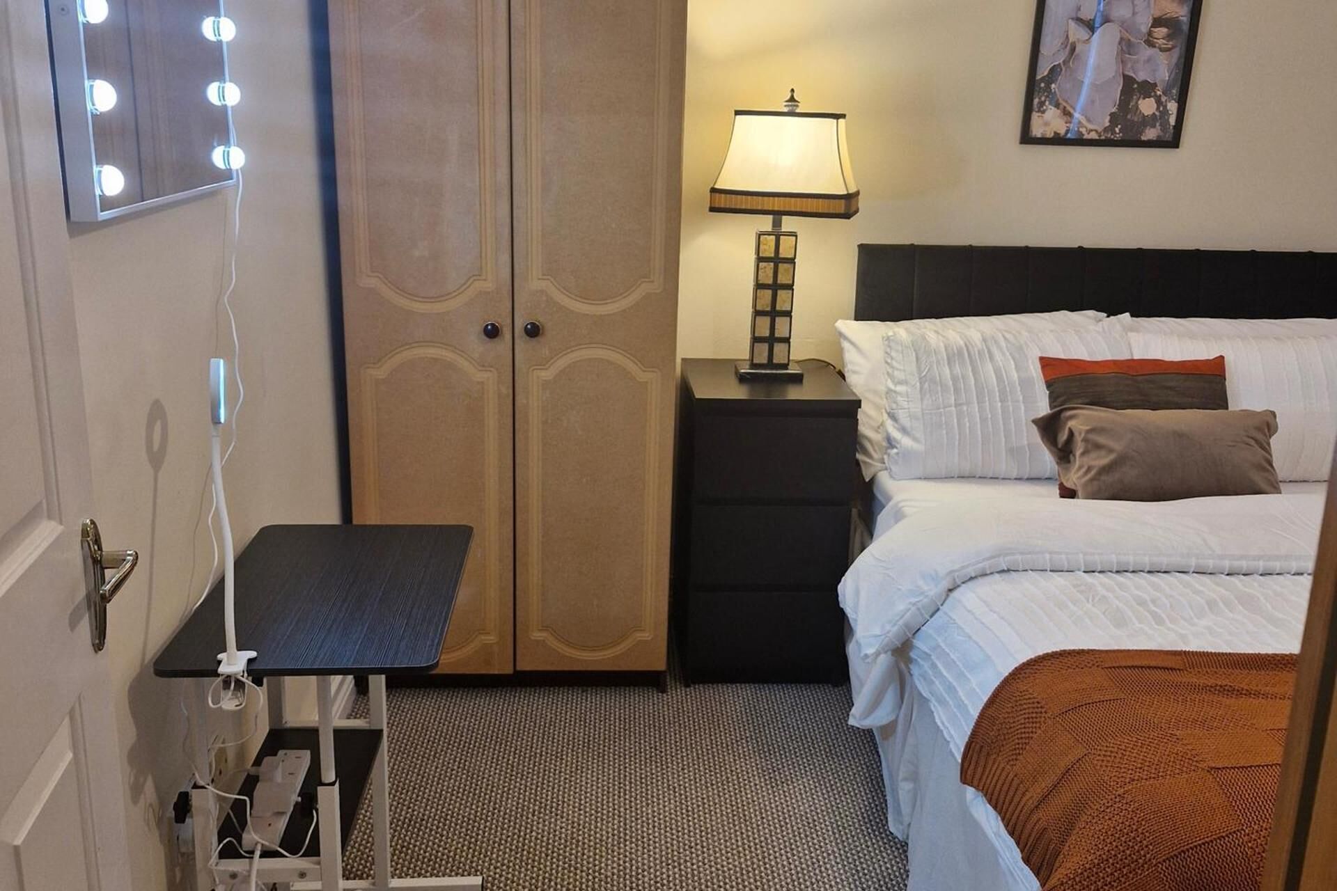A hotel room with a bed, bedside table, lamp, and a mirror with lights.