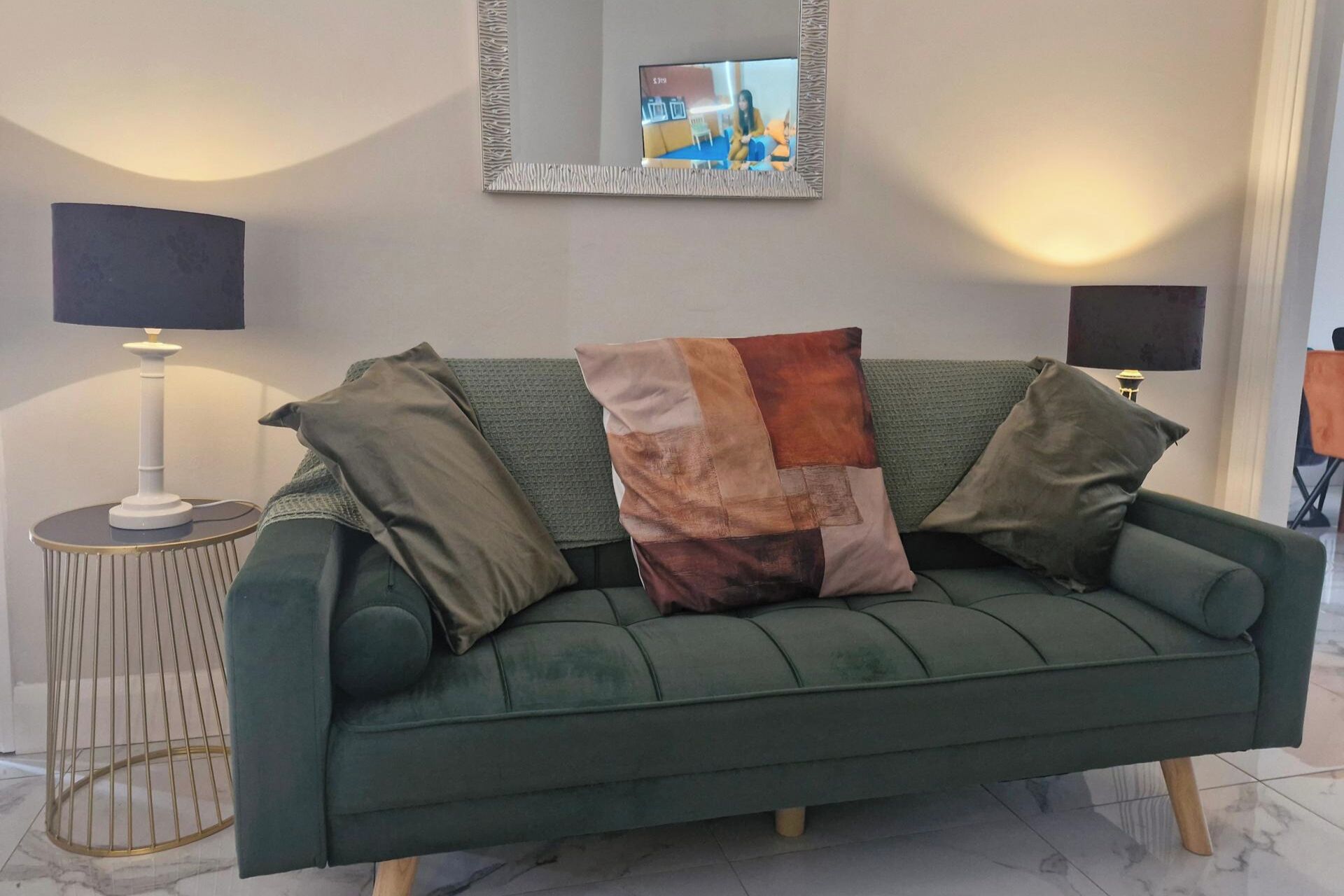 Ein grünes Sofa mit einem strukturierten Zierkissen, ein runder Beistelltisch mit Lampe und ein Spiegel an der Wand.