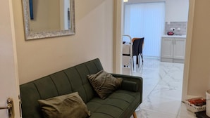 Un sofá verde con dos almohadas, un espejo, una zona de cocina con armarios y una mesa de comedor con sillas.