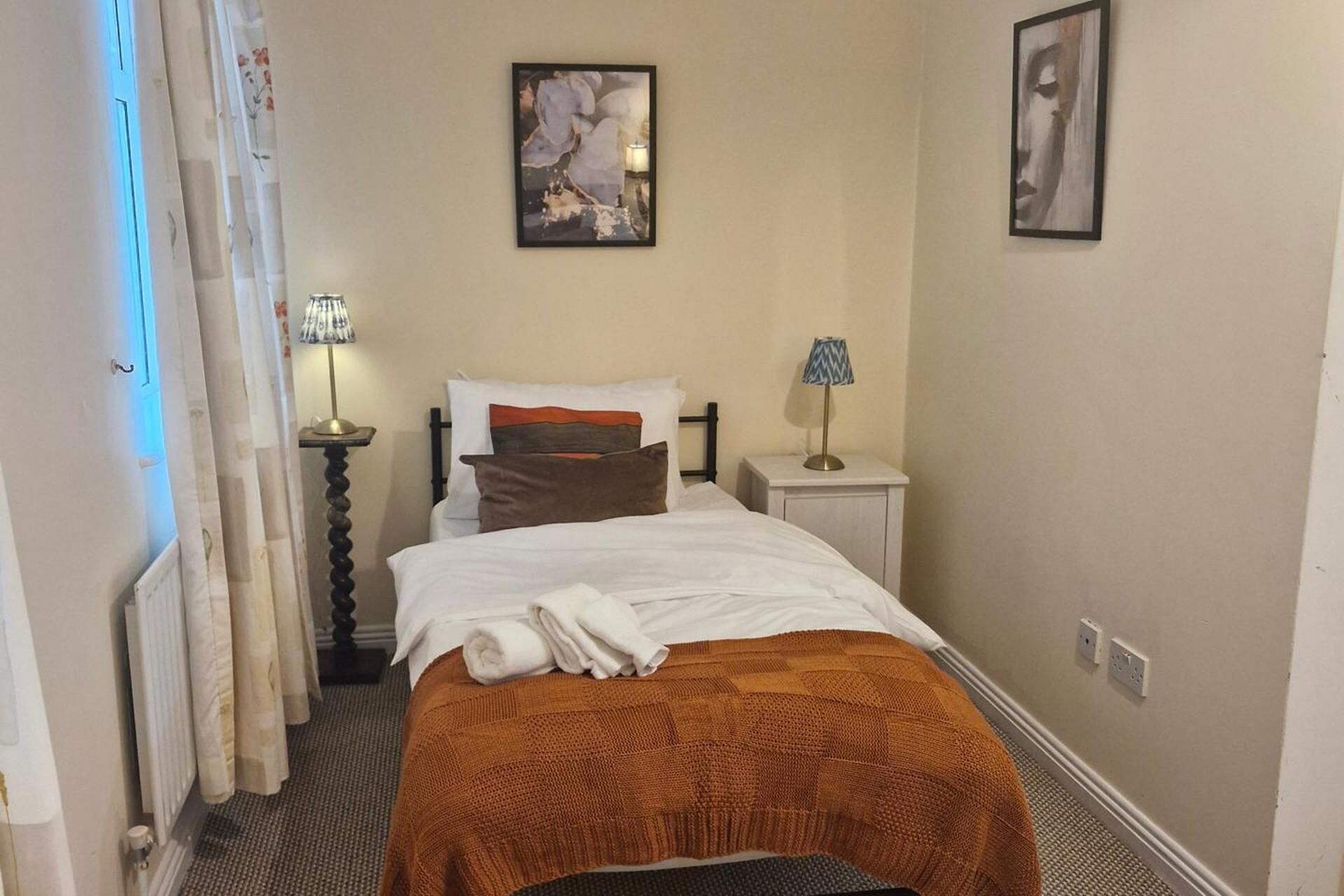 A hotel room with a bed, two bedside tables, a lamp, a radiator, and a curtain.