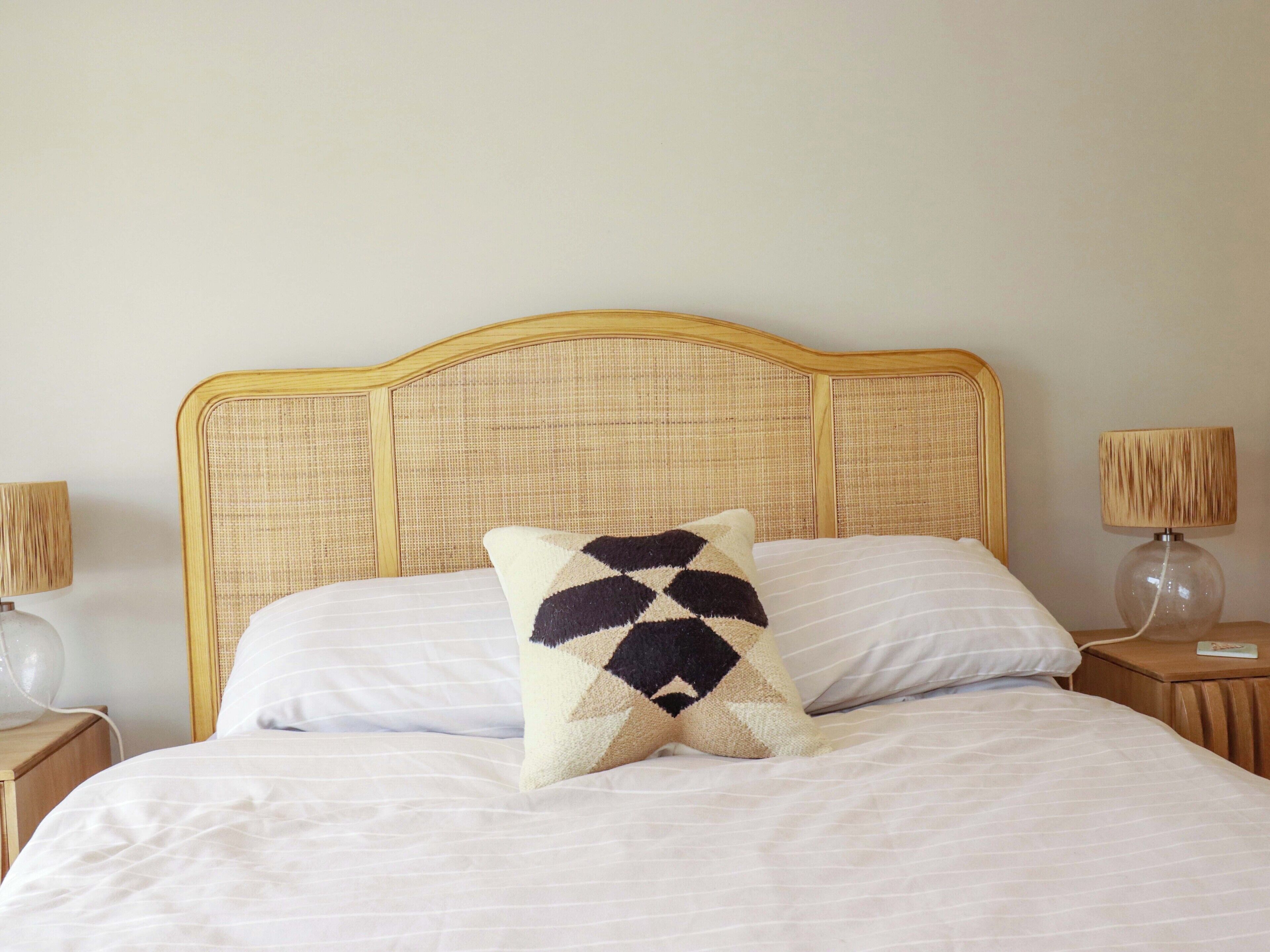 A bed with a geometric-patterned pillow, a plain white pillow, and a wooden headboard. There are two bedside lamps with lampshades on wooden nightstands.