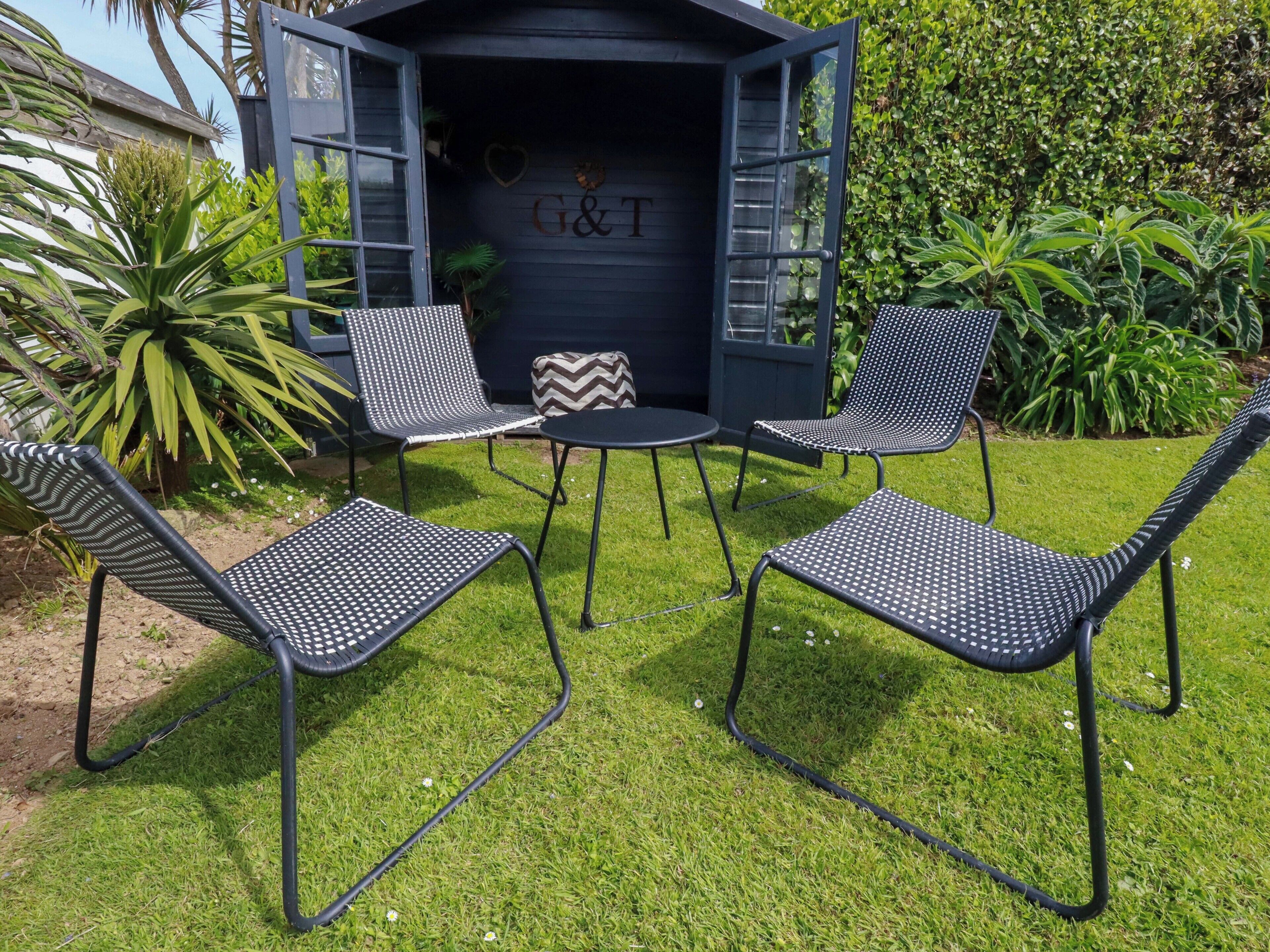An outdoor seating area with metal chairs and a table, surrounded by greenery and a dark-colored building with 'G&T' on the door.
