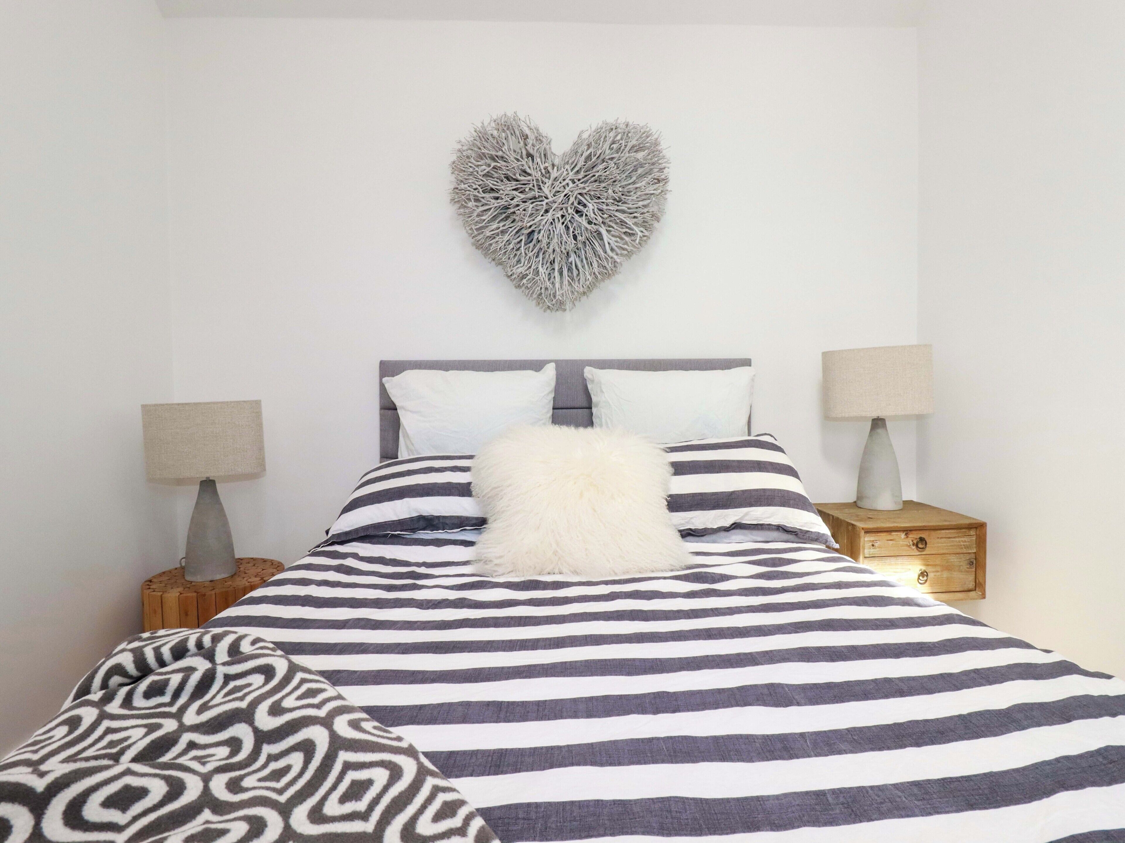 A bed with a striped bedspread, two pillows, a heart-shaped wall decoration, and two bedside tables with lamps.
