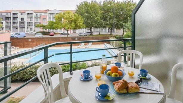 A balcony with a table set for breakfast, overlooking a swimming pool and a residential area. - Coastal Apartment Near Marina (Les Sables-d'Olonne)