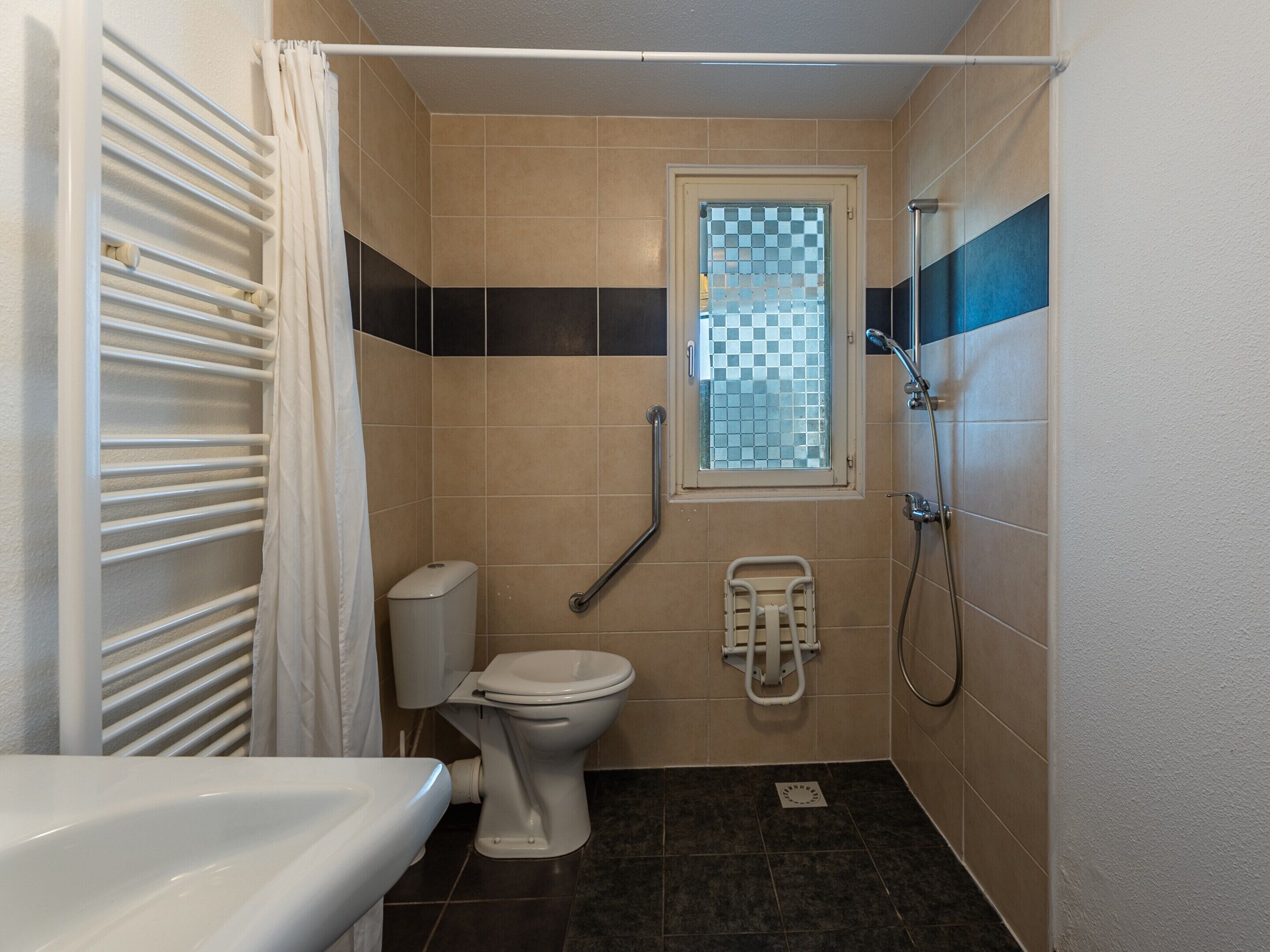 A small bathroom with a toilet, a shower, a window, and a handrail.