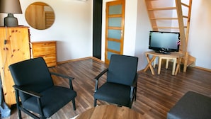 Habitació amb dues cadires negres, un armari de fusta, un mirall rodó, una televisió sobre un moble de fusta i una escala de fusta.