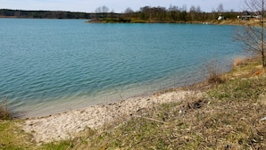 Un llac tranquil amb riba sorrenca, envoltat d'arbres i unes poques cases a la distància.