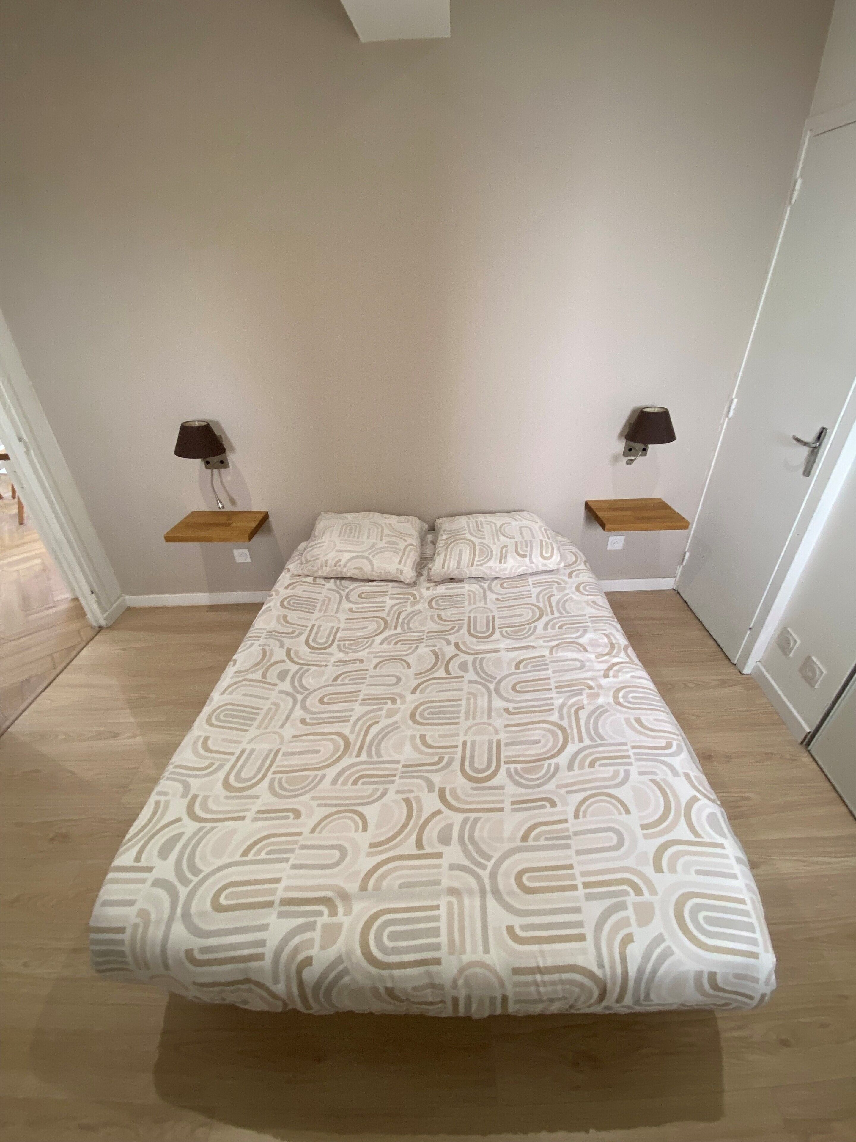 Une chambre à coucher comprenant un lit, deux tables de chevet, deux lampes fixées au mur et une porte.