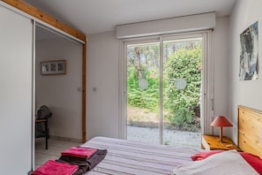 Un dormitorio con cama, mesita de noche, silla y una puerta corrediza de vidrio que da a un espacio exterior con vegetación.