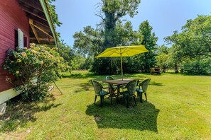 A red building with a covered porch, a table with chairs, and a yellow umbrella in a grassy yard.