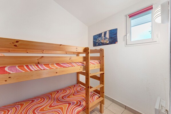 Una cama litera con estructura de madera y ropa de cama colorida, una ventana con vista al mar y un cuadro de un velero en la pared.