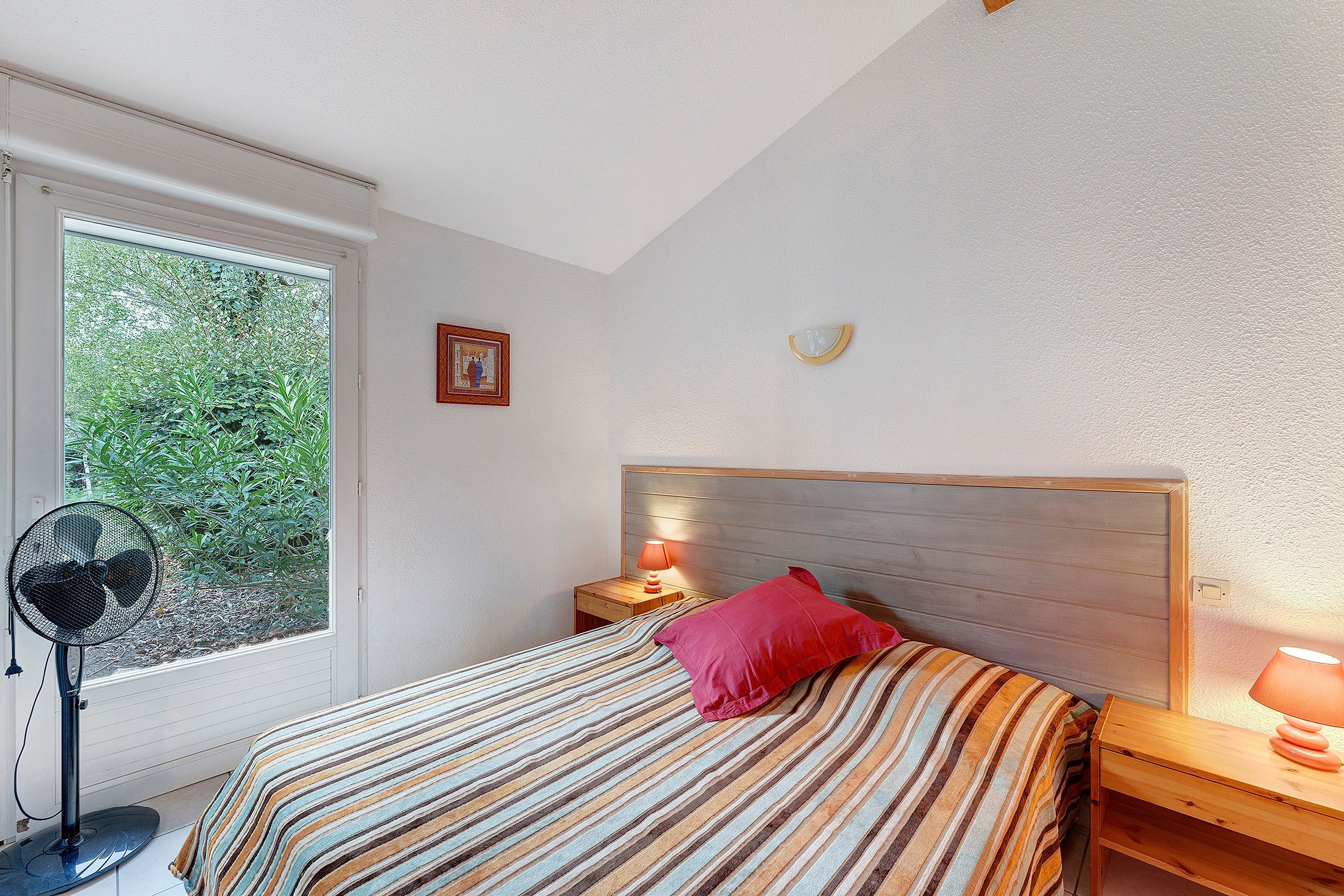 A bedroom with a bed, a nightstand, a lamp, a fan, and a window with a view of greenery.
