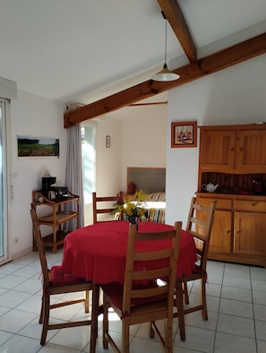 A dining area with a red tablecloth, wooden chairs, a hanging light fixture, and a kitchen area with wooden cabinets. - “4p Sanguinet - Addax” Apartment with Private Terrace and Wi-Fi (Sanguinet)