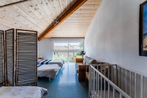A room with a sloped ceiling, a large window, two beds, a wicker sofa, and a crib.